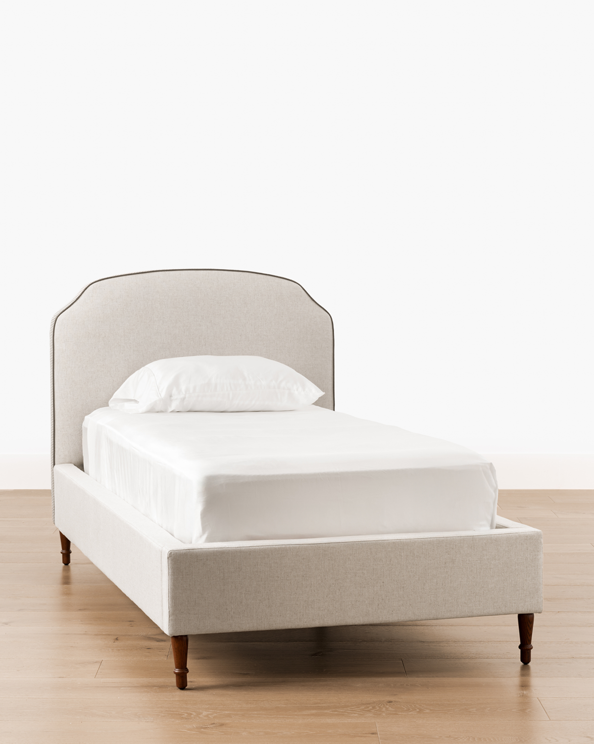 The Willoughby Bed features a light gray linen upholstered headboard and frame, white bedding, and English Oak legs, set on a light wood floor with a plain white background, by McGee & Co.