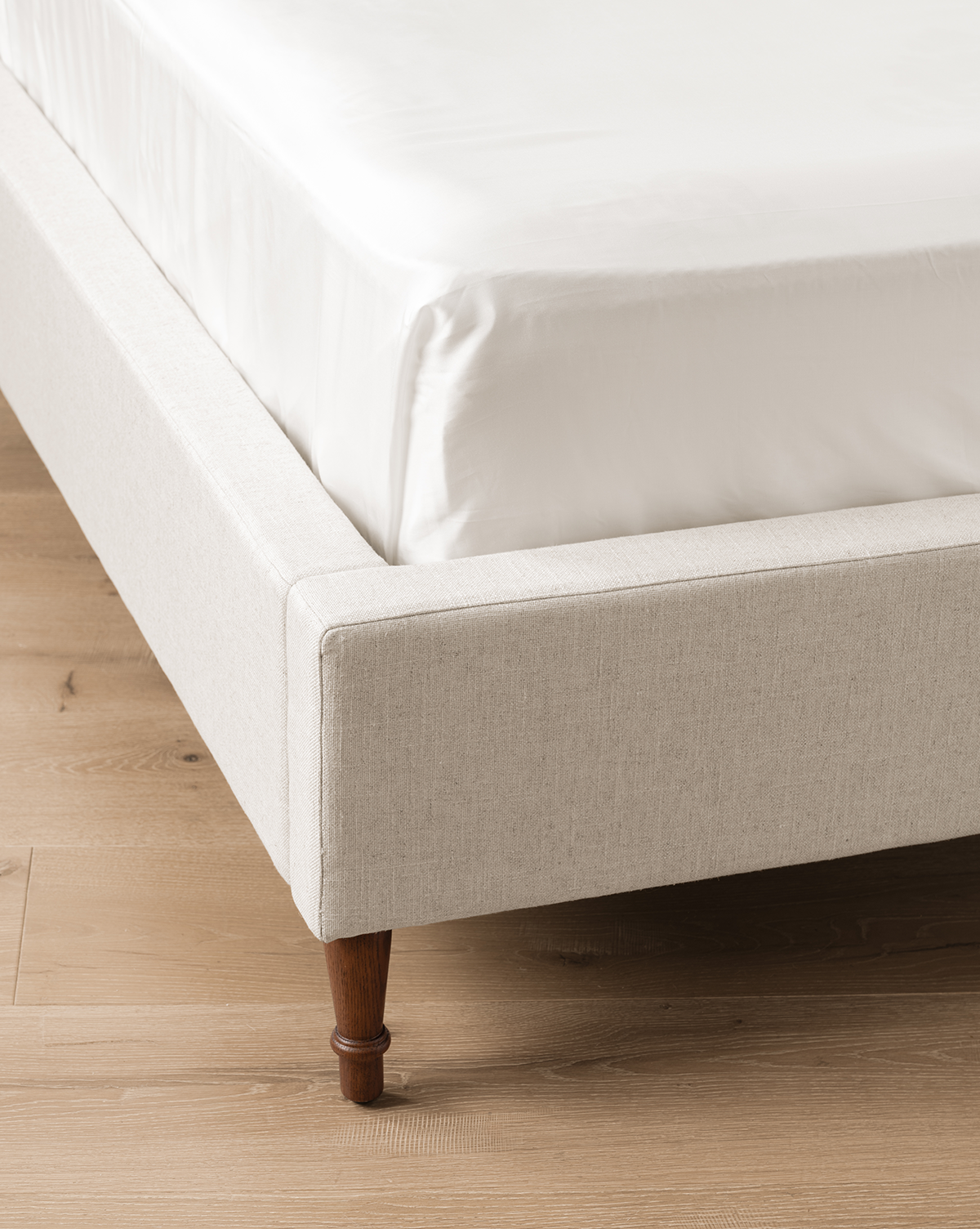 A close-up of the corner of the beige linen Willoughby Bed with English Oak legs, white mattress and bedding, set on a light wood floor, by McGee & Co.