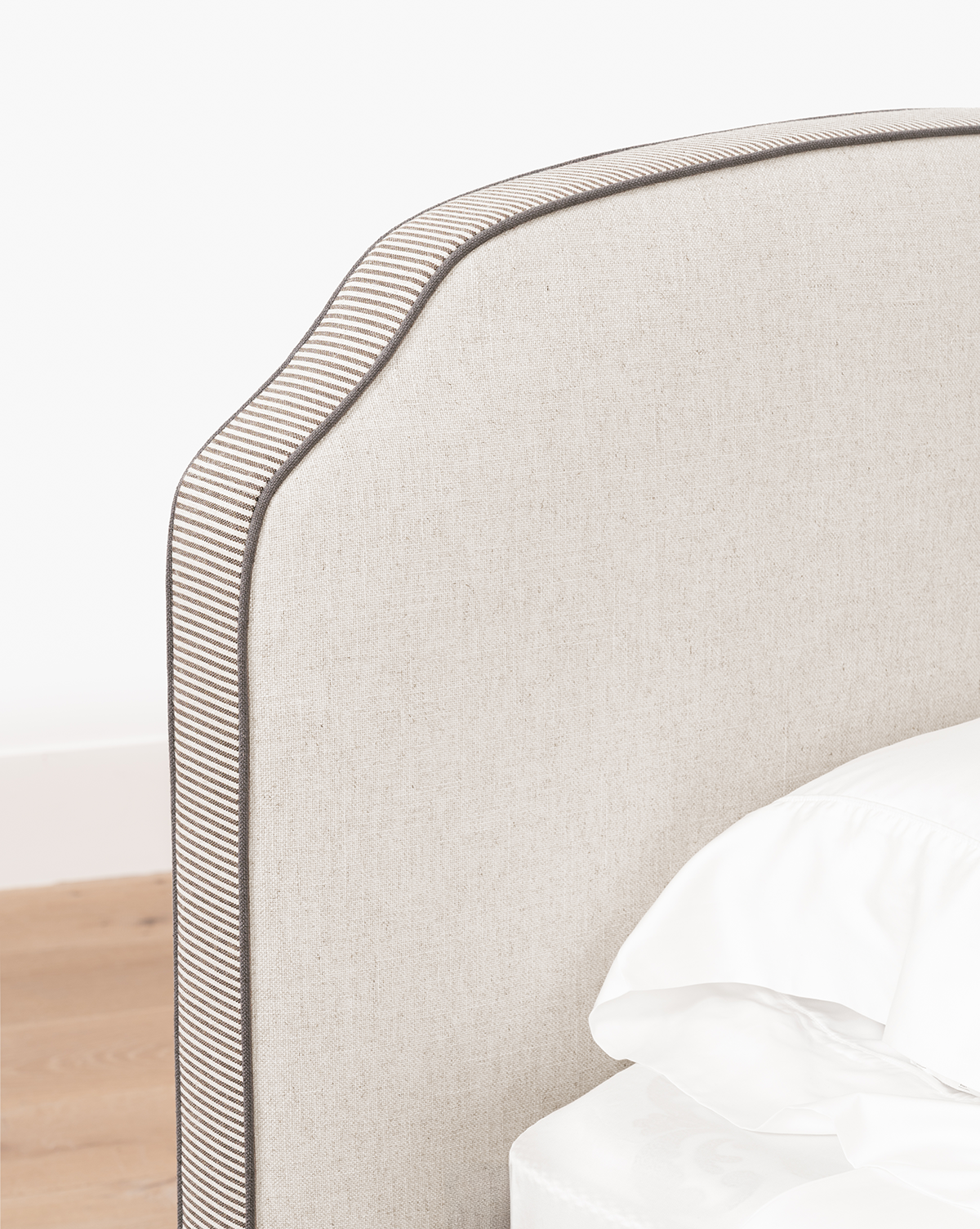 A close-up of the Willoughby Bed’s beige linen upholstered headboard, featuring a curved top and dark piping trim, shown beside white sheets and a light wood floor, by McGee & Co.