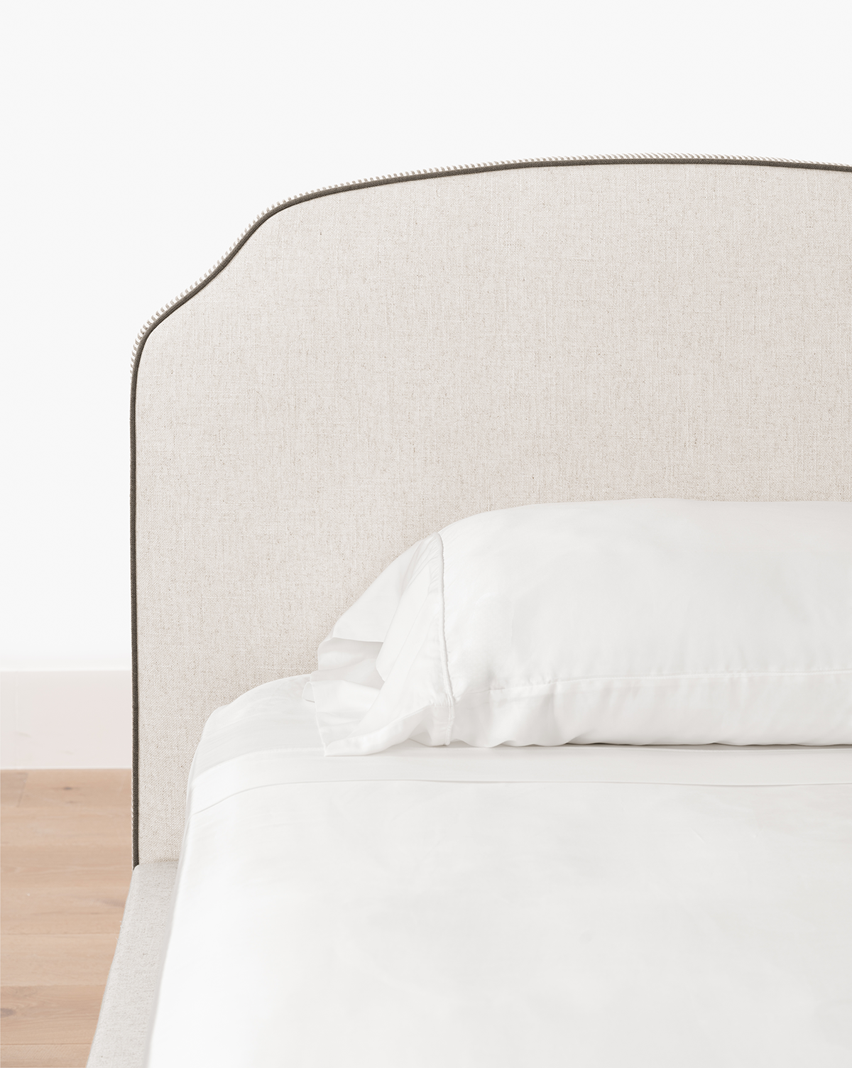The Willoughby Bed features linen upholstery, a light beige headboard, white pillows and bedding, elegant English Oak legs, set against a white wall and light wood floor, by McGee & Co.