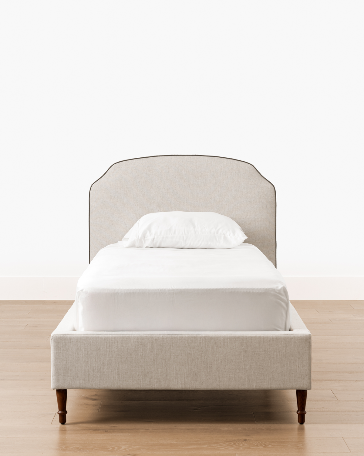 A Willoughby Bed with light beige linen upholstery, English Oak legs, white bedding, and a single pillow stands on a wooden floor against a plain white wall, by McGee & Co.