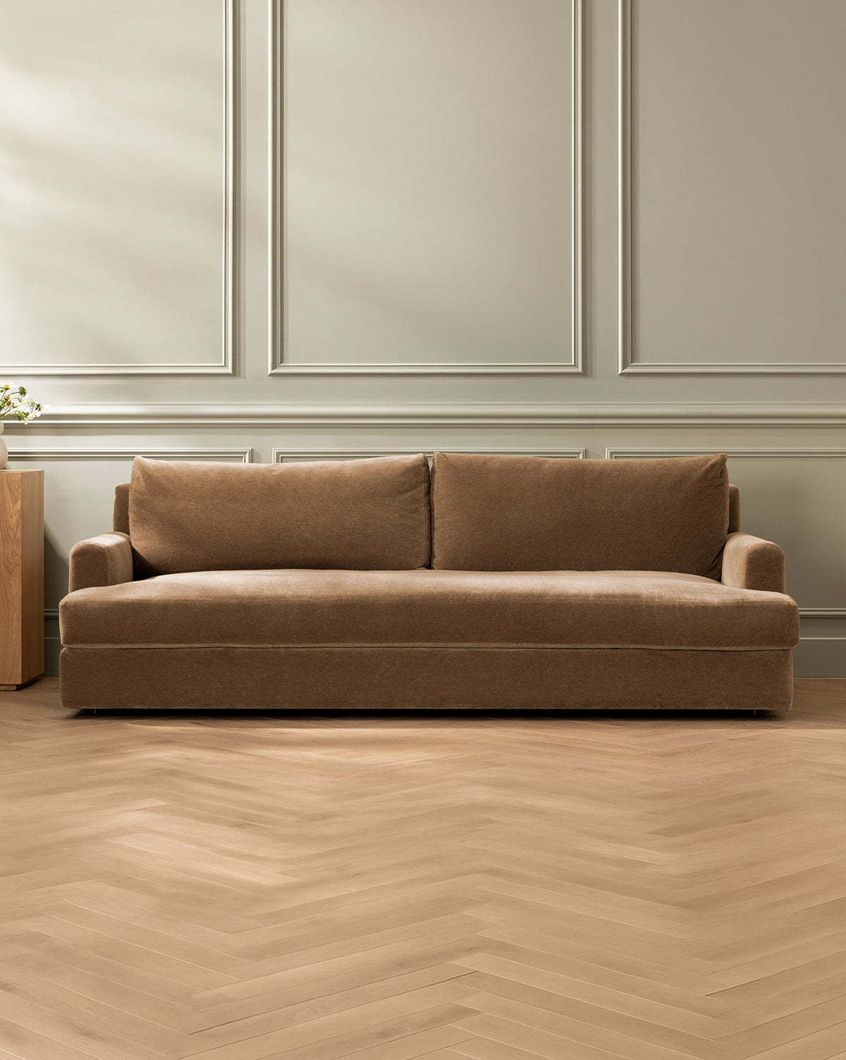 The Winthrop Sofa 90, upholstered in brown with two back cushions, sits in a minimal room with light gray paneled walls and light wood herringbone flooring, by McGee & Co. A small wooden side table is partially visible on the left.