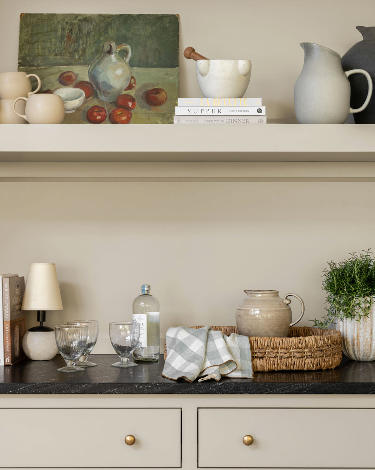 A kitchen countertop with glassware, a ceramic jug, folded checkered napkins, a potted plant, books, and a lamp. Above, a shelf displays pottery—including the Brown Stoneware Pitcher—books, and a still life painting with fruit and a white pitcher by McGee & Co.