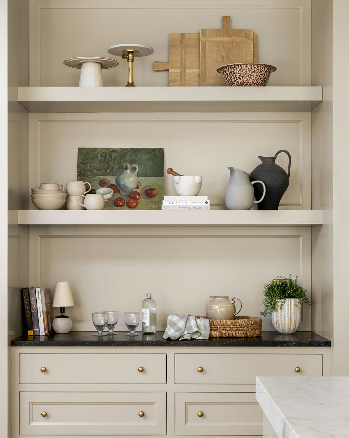 Open shelves with neutral dishware, glassware, a Rectangle Bread Board in a natural finish, oak and walnut cutting boards, a plant, lamp, books, bottle, and a small framed fruit painting—all atop cream cabinetry with gold knobs by McGee & Co.