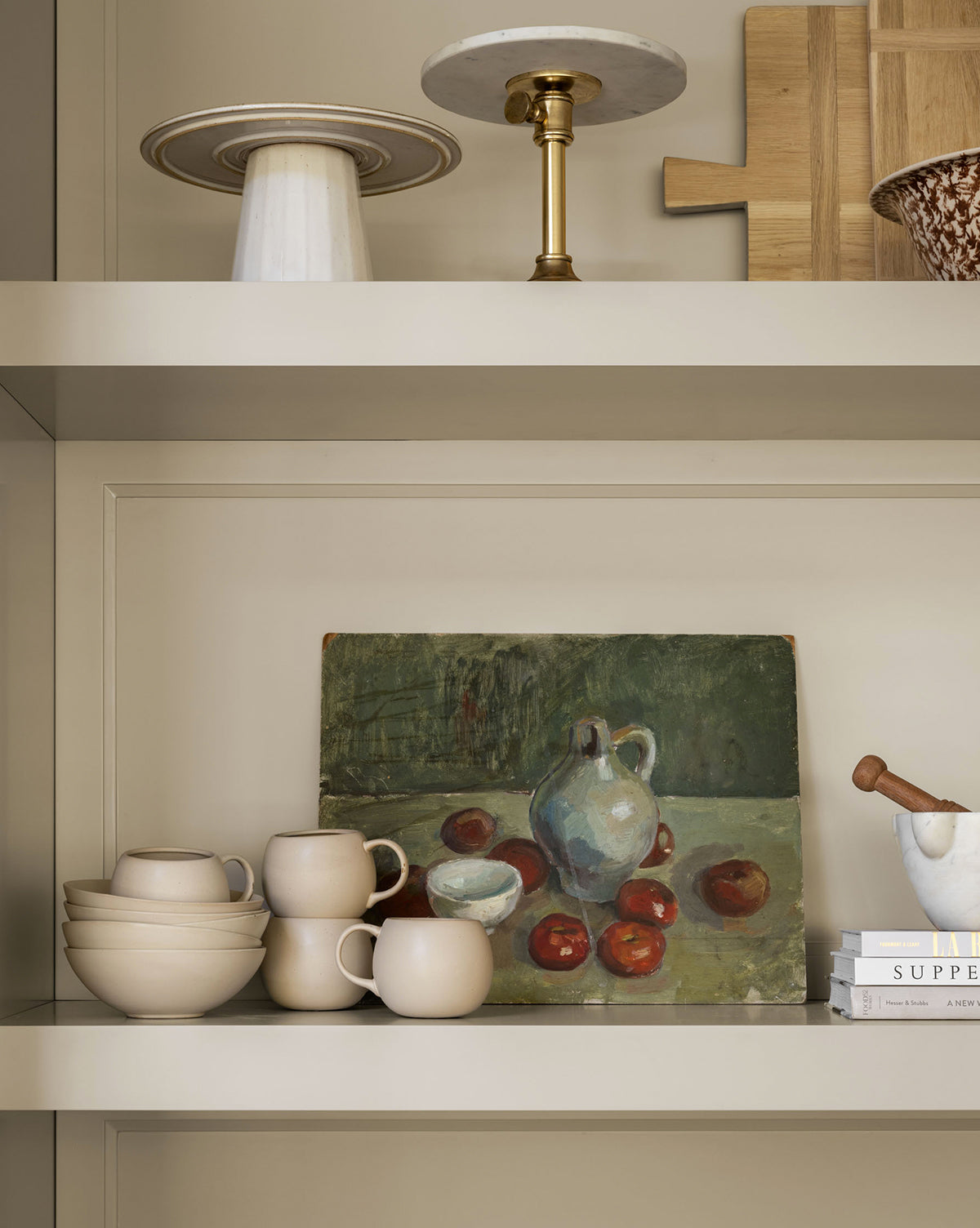 Neutral shelves display glazed stoneware, including the Luana Bowl, cups, a marble cake stand, cutting boards, books, a mortar and pestle, and a still-life painting—creating a modern space with neutral charm, by McGee & Co.