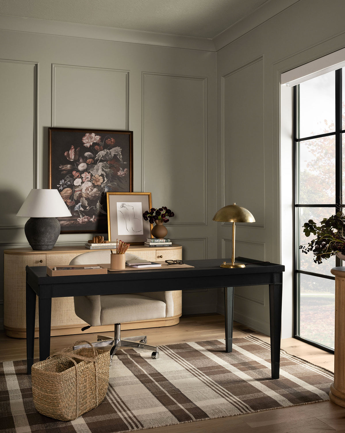 A modern home office with a black desk, beige chair, two lamps, framed floral art, woven basket, and large window. The paneled walls add texture, while the floor features the Orton Handwoven Wool Rug in a plaid pattern by McGee & Co.