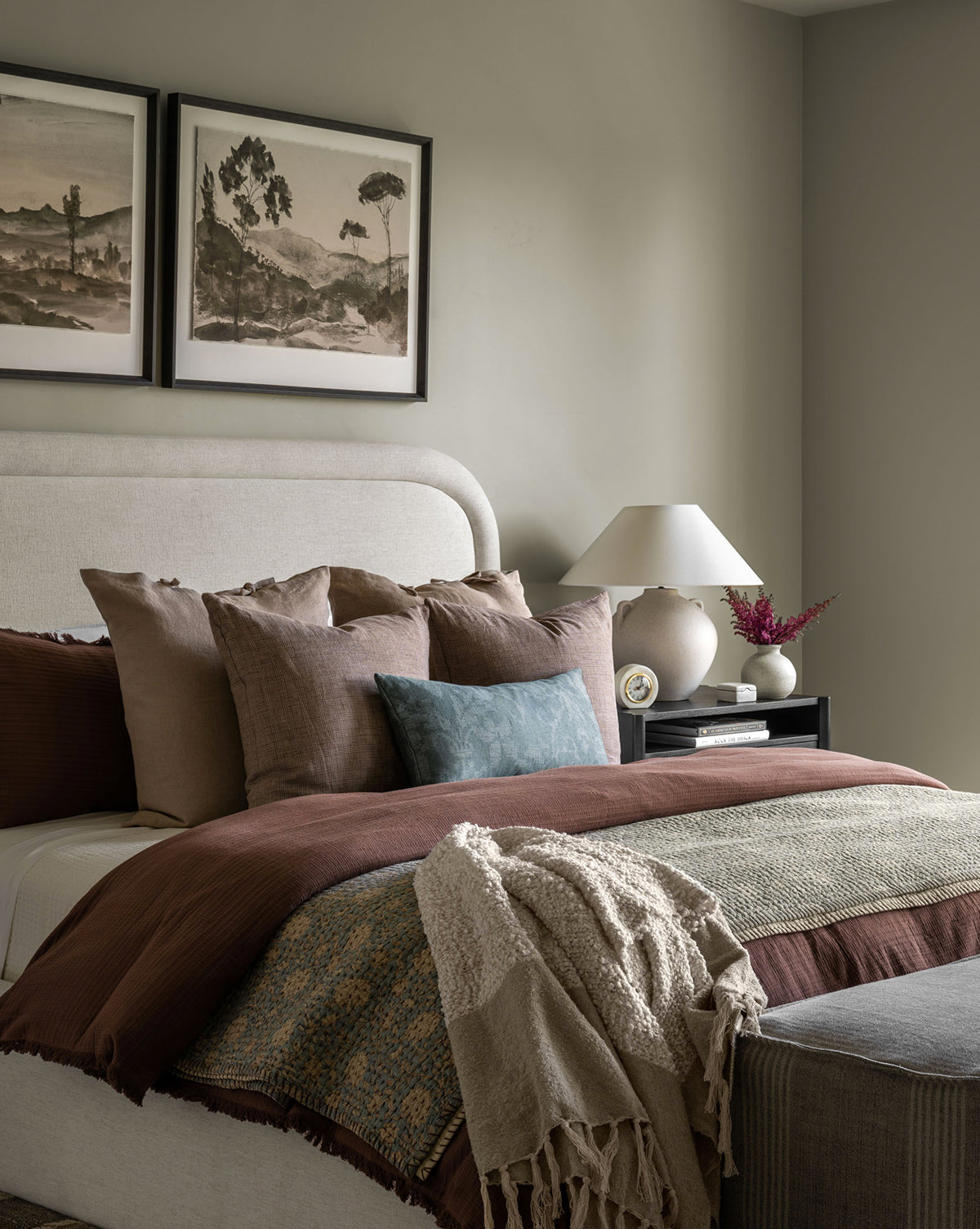 A neatly made bed features the Gabriella Block Print Quilt and layered pillows in earthy tones, next to a black nightstand with a lamp, clock, and vase. Two landscape artworks hang above the upholstered headboard.