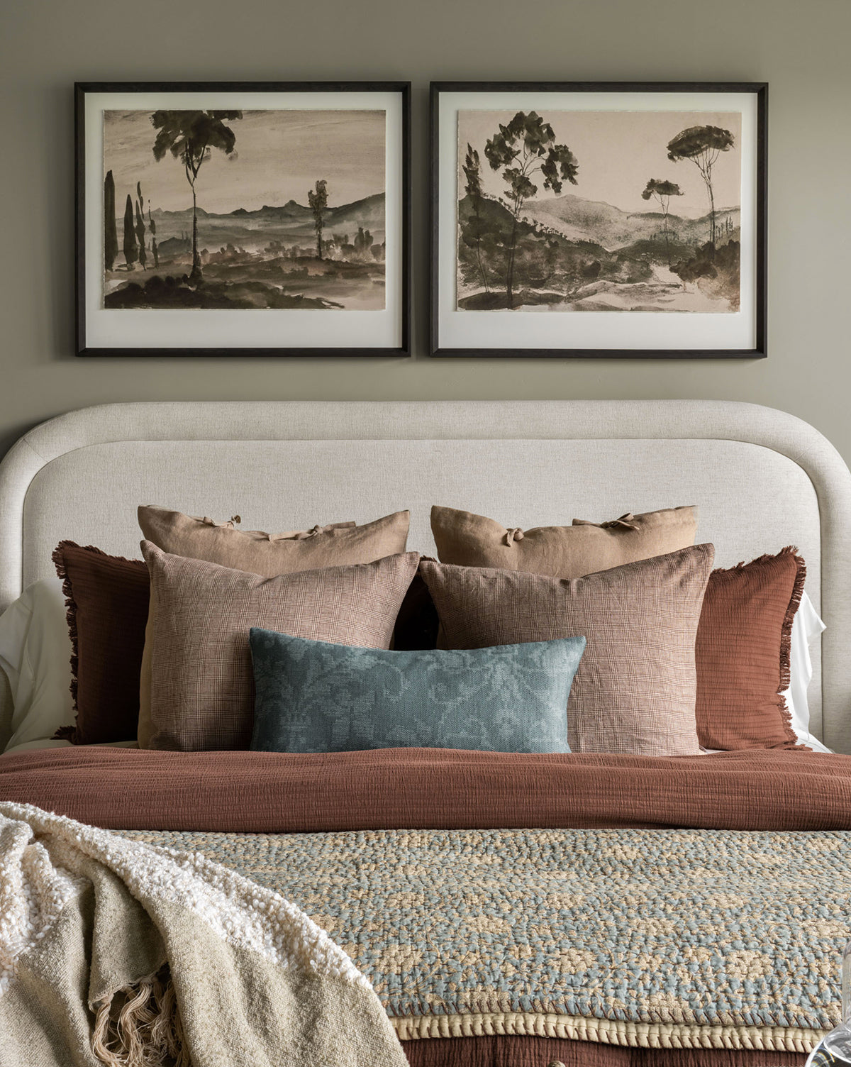 A neatly made bed with neutral, earthy-toned pillows and blankets sits against a light wall. Above the bed, the Washed Terrain I watercolor art print in a black frame adds a calm, natural touch to the bedroom decor.