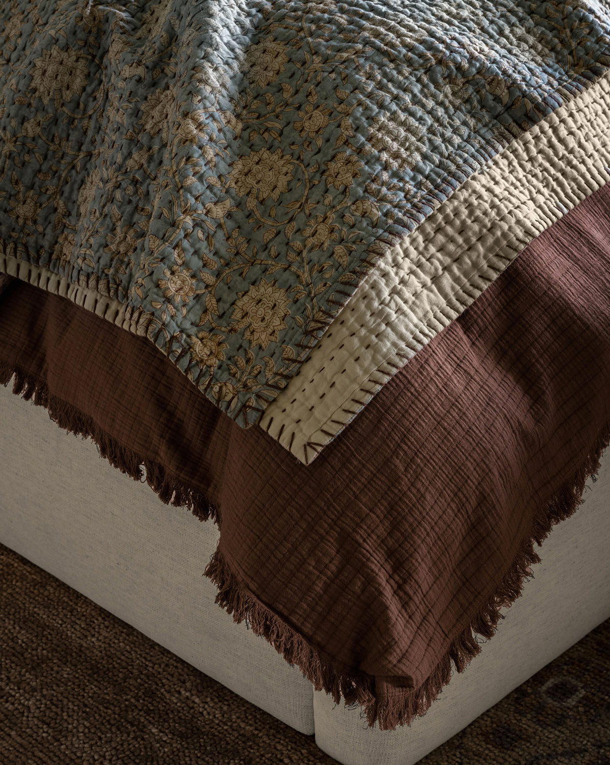 A close-up of layered bedding on a bed, featuring a textured blue and beige quilt over the Hendricks Chocolate Cotton Duvet Cover and a brown fringed blanket on a light-colored upholstered base.