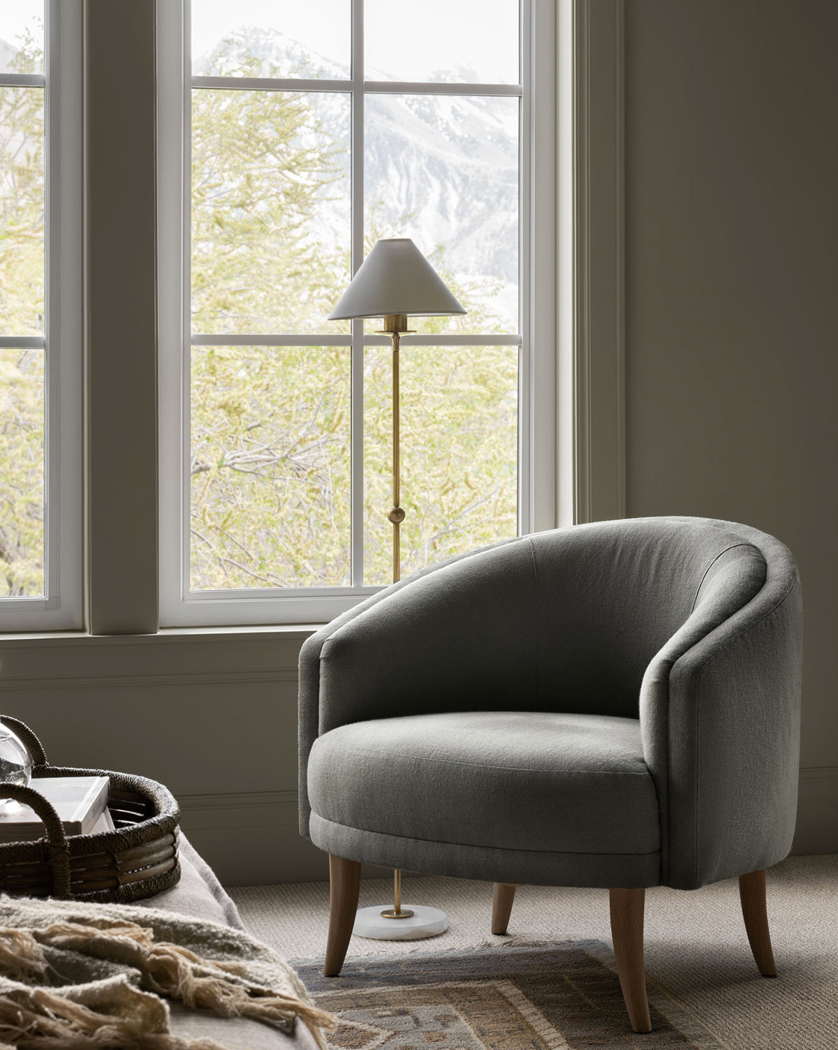 The cozy Marguerite Lounge accent chair in gray sits by a large window with mountain and tree views, paired with a brass floor lamp and a woven basket nearby. McGee & Co.