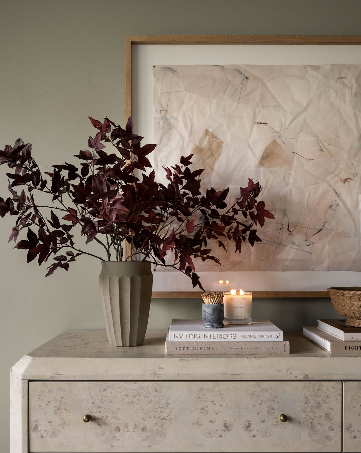 A beige stone console table featuring the Frida Vase with burgundy leaves, stacked design books, lit candles, and a bowl, arranged beneath an abstract artwork on a muted wall.
