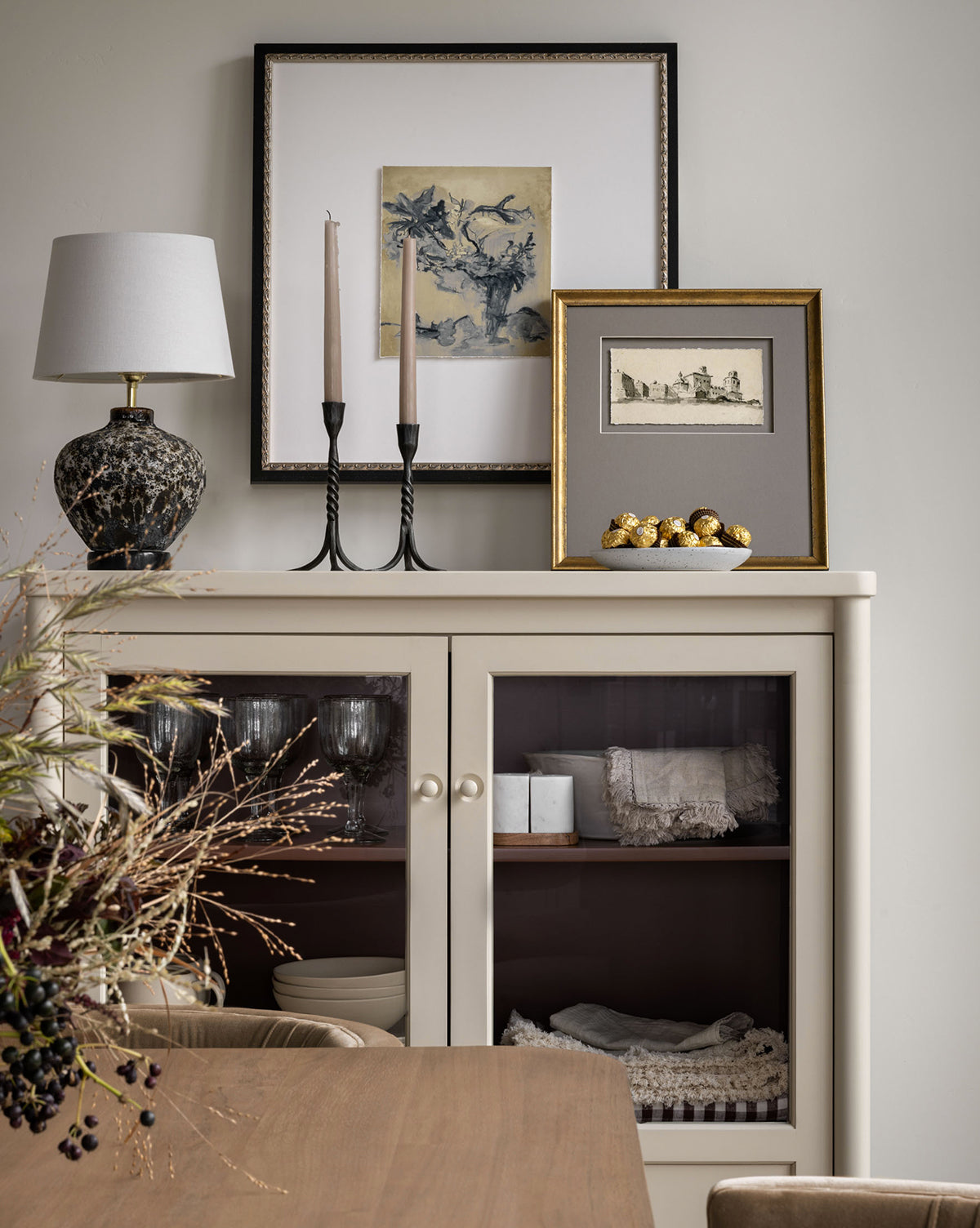 A decorated cabinet with glass doors displays linens and glassware. On top are a lamp, two candlesticks, a bowl of golden ornaments, and the Sepia City artwork against a neutral wall, with a dried floral arrangement nearby.
