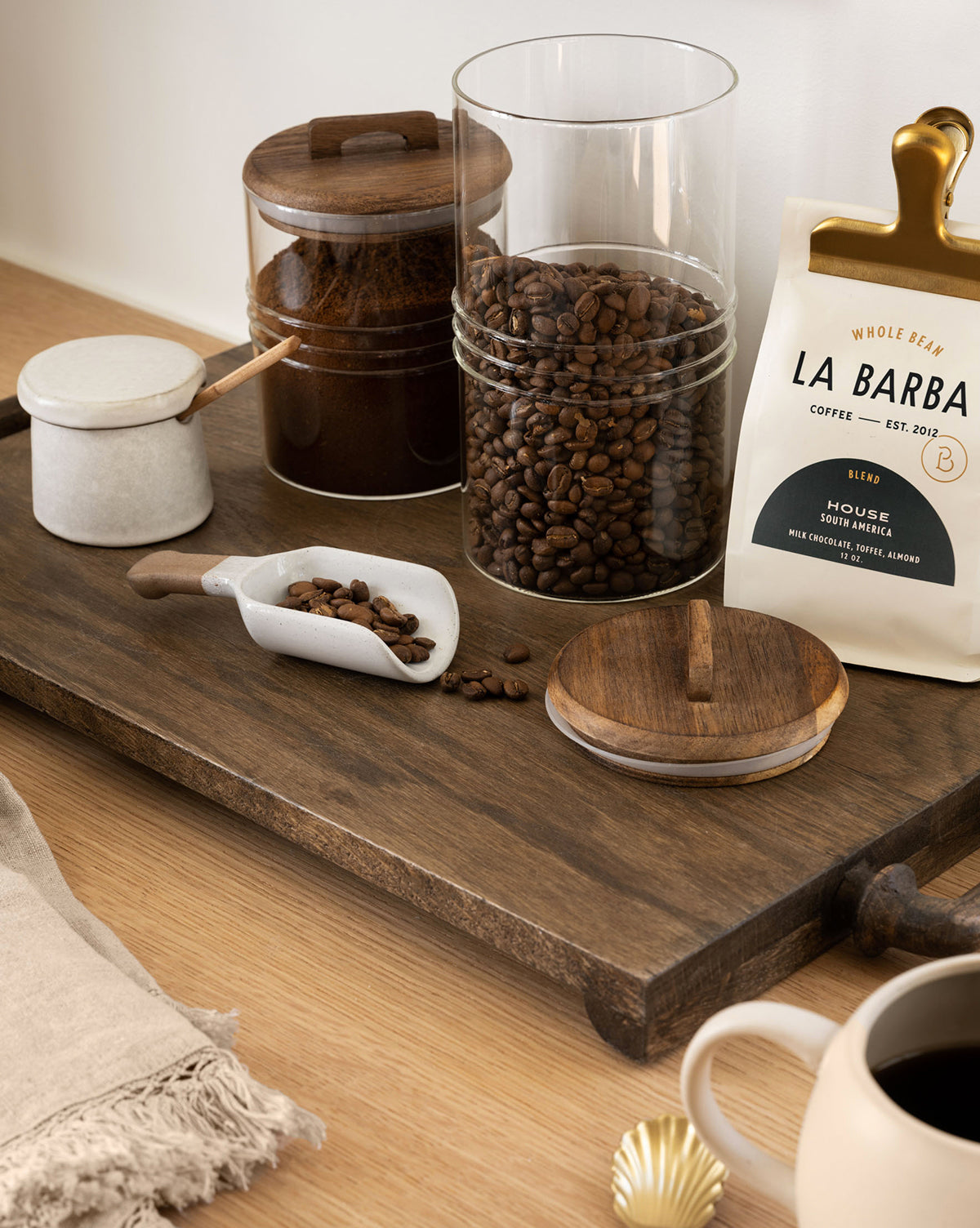 The Fordham Board displays coffee beans in glass jars, ground coffee in a jar, a “La Barba Coffee” bag, a ceramic scoop with beans, a cup of black coffee, and a linen napkin on a wooden surface, by McGee & Co.