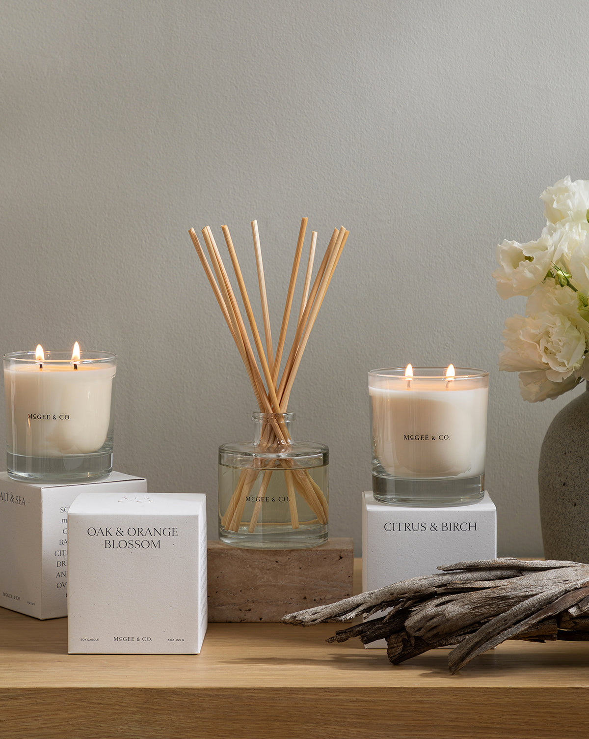 A minimalist display with two lit soy candles, a glass diffuser, white flowers in a vase, driftwood, and luxury candle boxes—including one labeled Citrus & Birch Candle—all arranged on a light wooden surface, by McGee & Co.