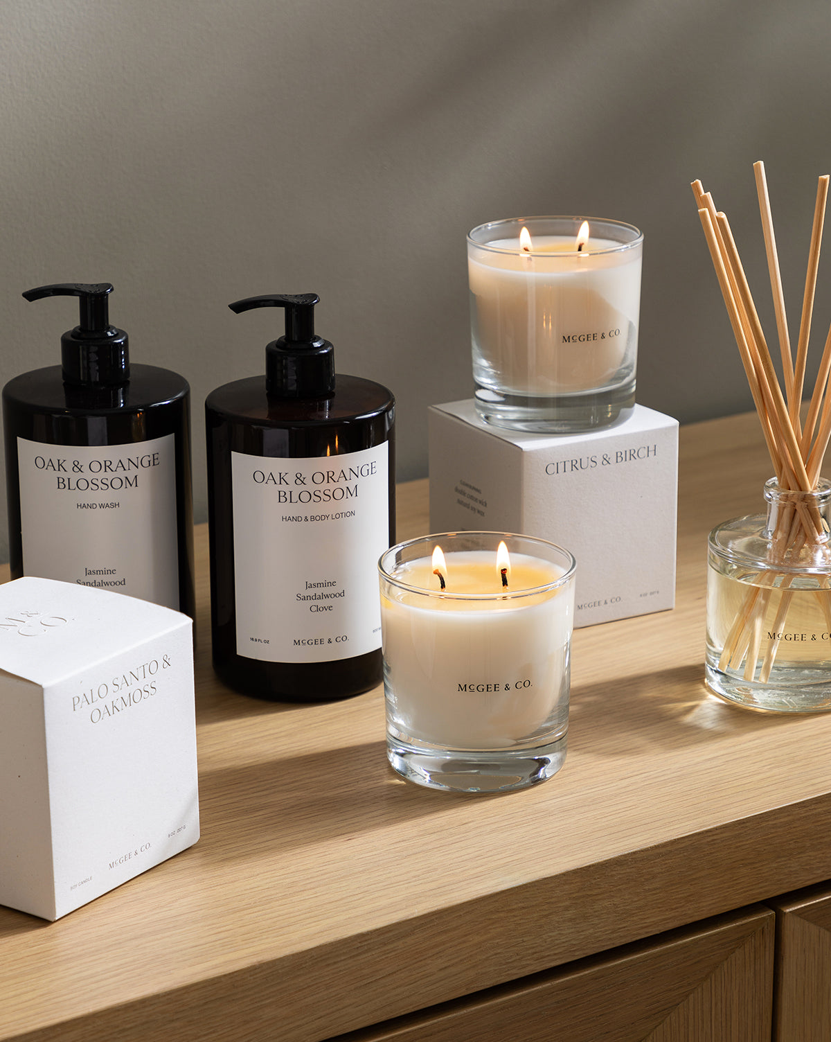 A wooden surface displays the minimalist white and black Palo Santo & Oakmoss Candle in a lit glass jar, along with pump bottles, another candle, boxes labeled Citrus & Birch and Palo Santo & Oakmoss, and a reed diffuser by McGee & Co.