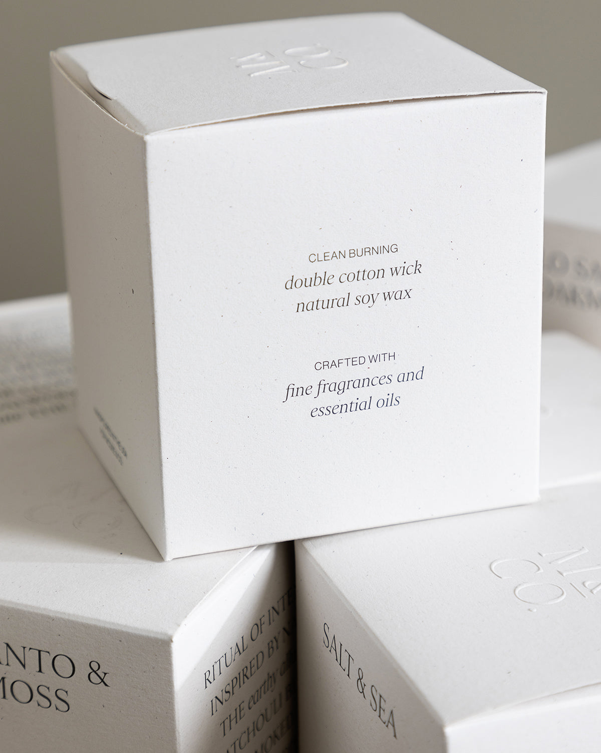 A white candle box with minimalist text describes the Palo Santo & Oakmoss Candle—clean burning, double cotton wick, crafted with fine fragrances and essential oils. Similar boxes, like an essential oil candle, are partially visible underneath by McGee & Co.