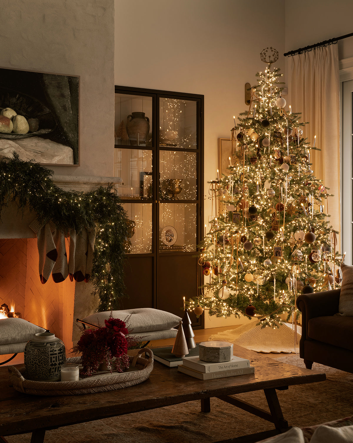 A warmly lit living room decorated for Christmas features a glowing 9' Grandis Fir Pre-Lit Faux Tree with ornaments, stockings on a garlanded fireplace, and cozy cushions with books atop a wooden coffee table, by McGee & Co.