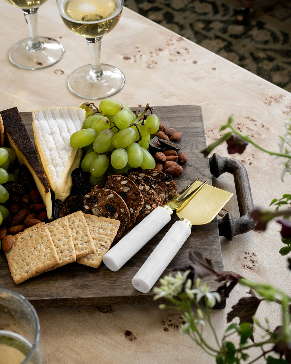 The Fordham Board, crafted from solid oak, beautifully presents cheese, green grapes, crackers, nuts, and seeded crisps. A marble-handled cheese knife and fork accompany it, with two glasses of white wine placed nearby by McGee & Co.