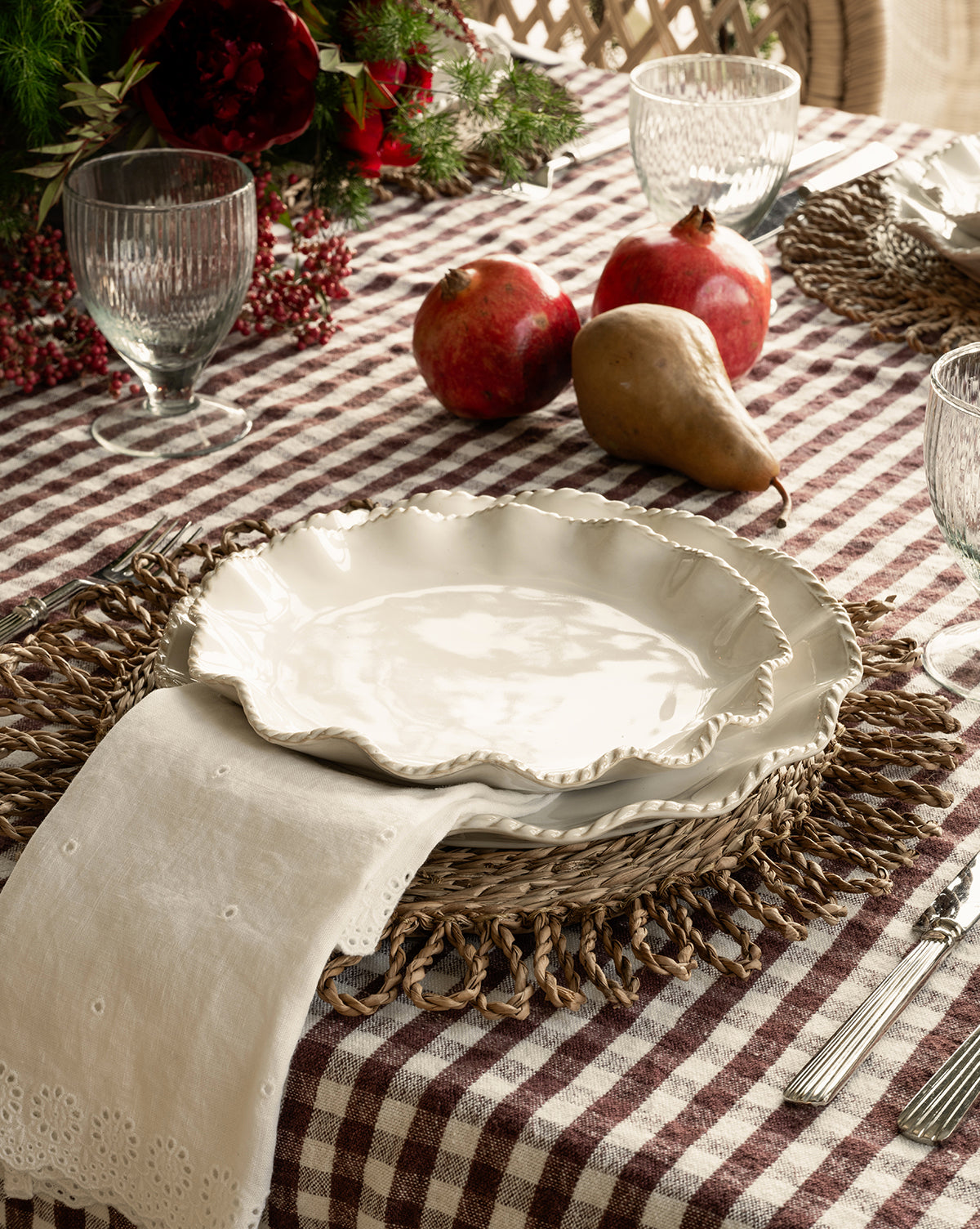 A rustic table with white ceramic plates on woven chargers, a checkered cloth, glassware, silverware, two pomegranates, a pear, greenery with red berries, and McGee & Co. x Loeffler Randall Delilah Broderie Napkins by McGee & Co.