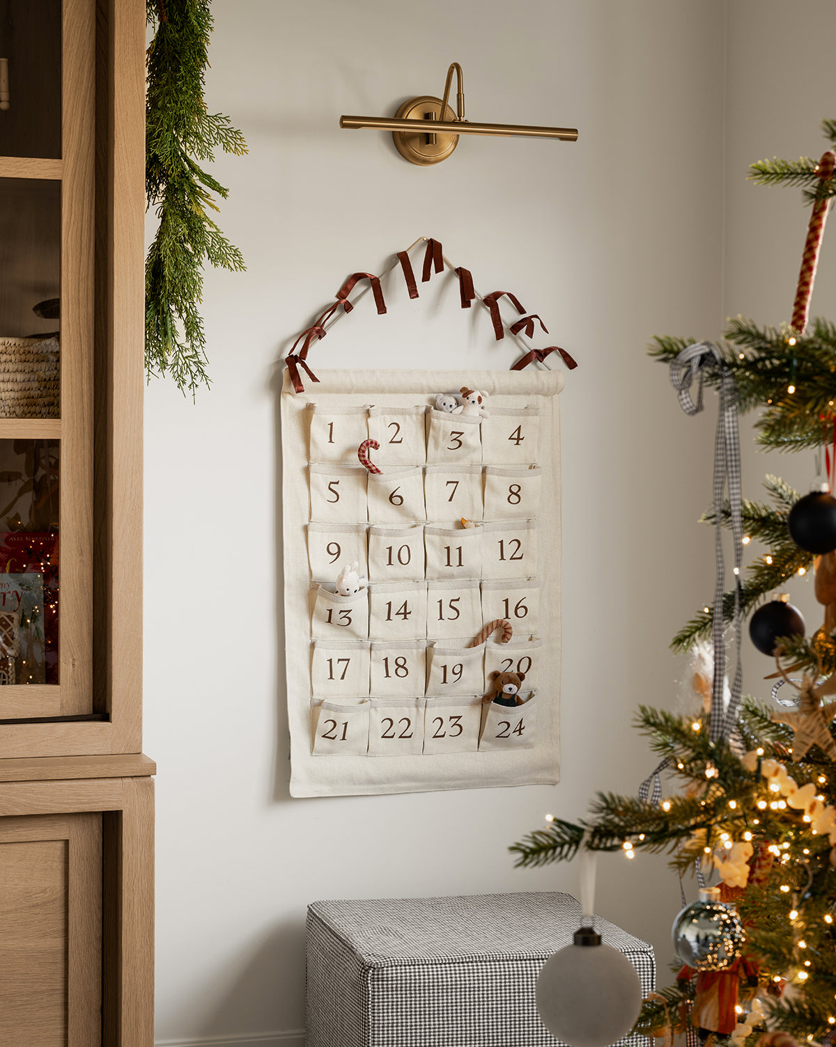 An Advent Calendar with numbered pockets hangs on the wall, decorated with small gifts. Nearby, a lit Christmas tree and festive greenery on a cabinet create a cozy holiday atmosphere, by McGee & Co.