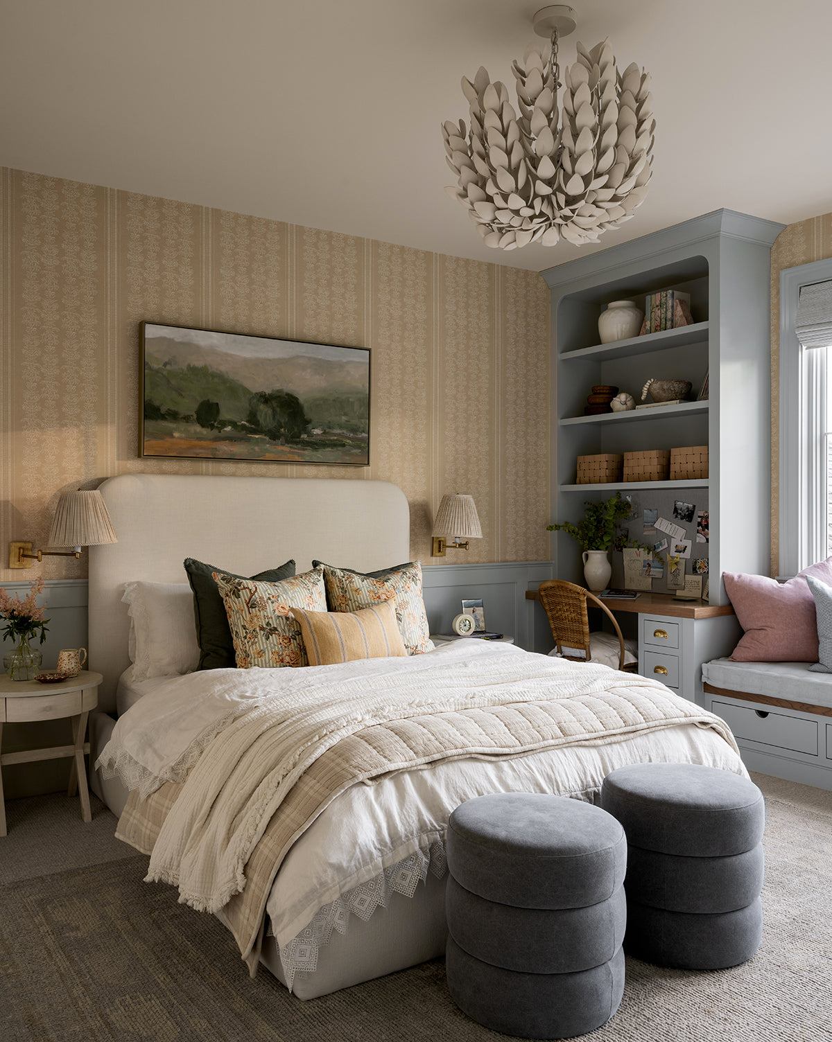 A cozy bedroom featuring the Northcott Bed layered in soft bedding and throw pillows, two round gray ottomans at the foot, built-in shelves, a window seat, a desk with chair, and a leafy chandelier overhead, by McGee & Co.