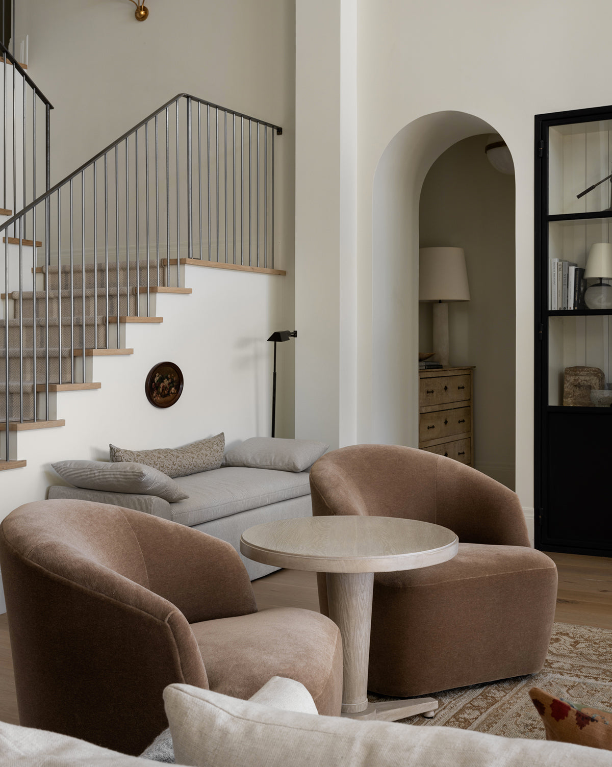 A modern living room features two Makerspalm Byron Chairs in brown paired with a round table, a light gray sofa by the stairs, neutral decor, and a black-framed glass cabinet beside an arched doorway—all thoughtfully styled in collaboration with McGee & Co.