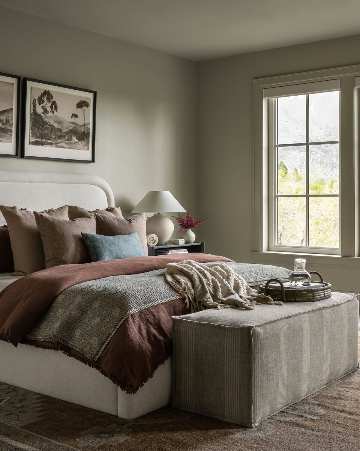 A cozy bedroom features a beige upholstered bed with brown and blue bedding, decorative throw pillows, and a Johannes Bench at the foot holding a tray with a glass carafe. A bedside table, lamp, and window complete the inviting scene, by McGee & Co.