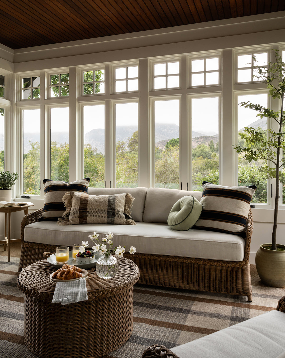 A cozy sunroom with large windows, a wicker sofa with striped pillows, the weather-resistant Haviland Coffee Table topped with drinks and pastries, potted plants, and views of trees and mountains in the background, by McGee & Co.