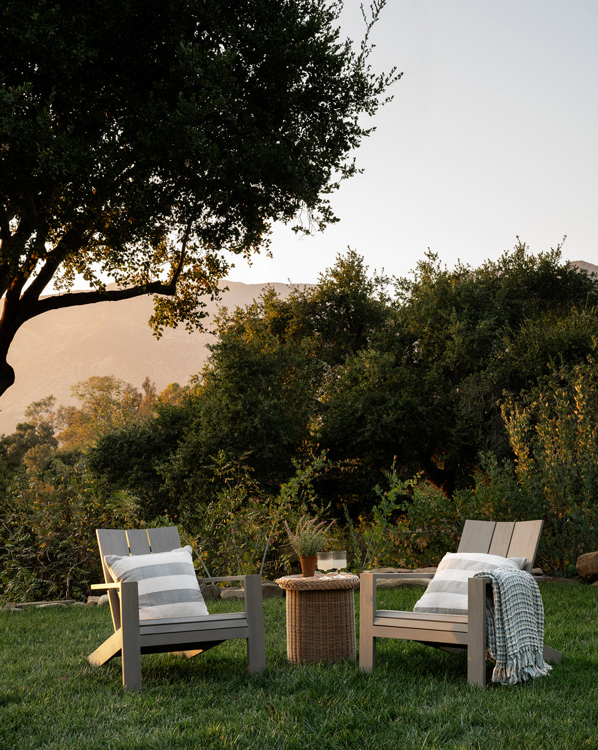 Two Triby Gray Teak Outdoor Lounge Chairs with cushions and throws sit on grass facing a small round wicker table with drinks and a plant, surrounded by greenery and trees, with mountains visible in the sunset—a relaxing outdoor setup, by McGee & Co.