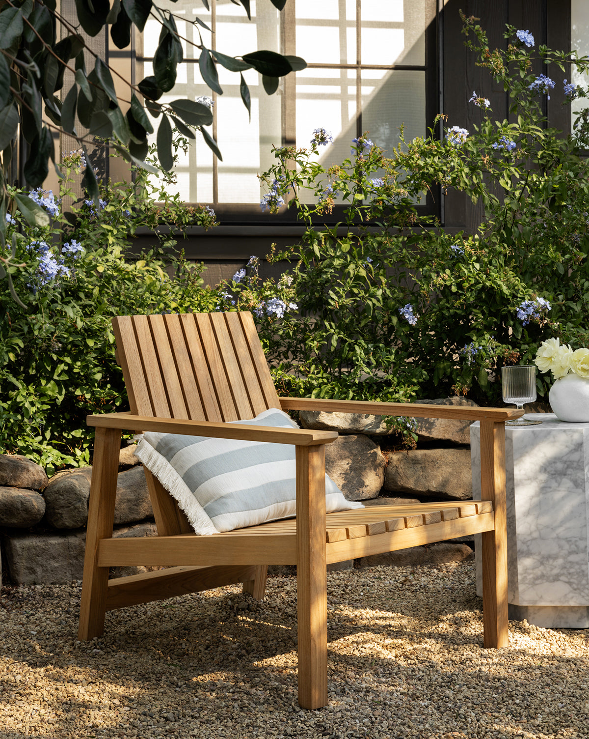 The Gilda Outdoor Lounge Chair with a striped cushion rests on gravel beside a small marble side table with flowers, set against lush green bushes and blooming purple flowers in front of a house’s window, by McGee & Co.