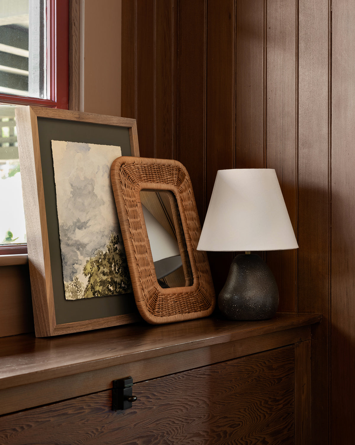 A framed painting, a wicker-framed mirror, and the Vedruna Table Lamp with a tapered white shade sit on a wooden chest against wood-paneled walls near a window, by McGee & Co.