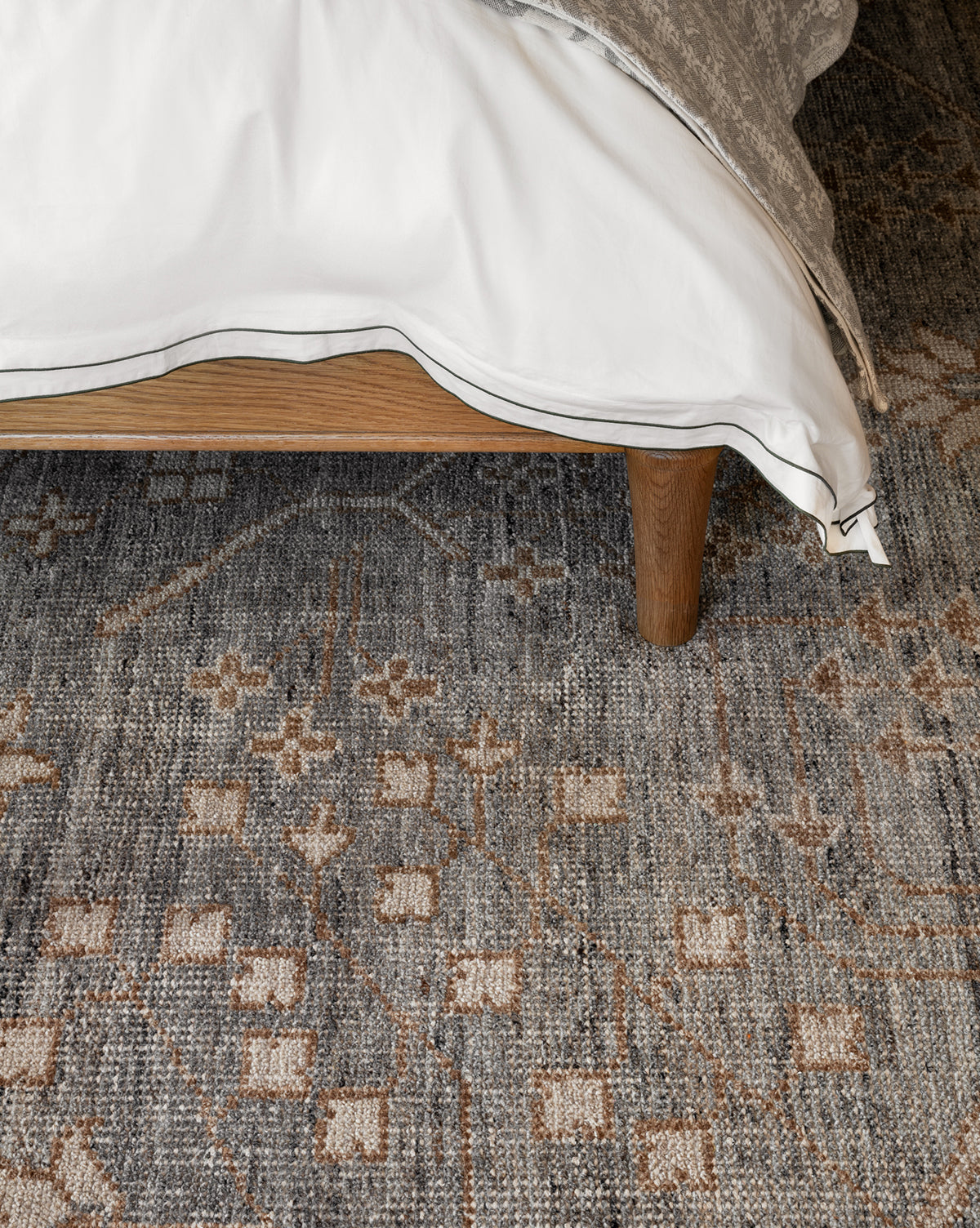 A corner of a bed with white and beige bedding sits on a wooden frame above the Elison Hand-Knotted Wool Rug, featuring geometric and floral motifs that highlight artisan craftsmanship, by McGee & Co.
