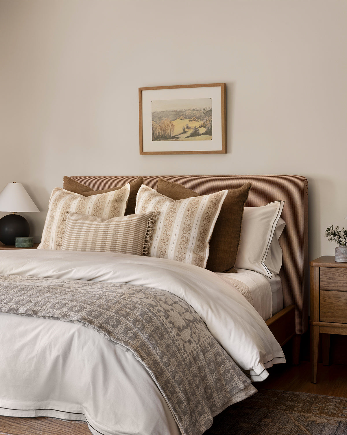 A cozy bedroom features a beige upholstered headboard, neatly arranged neutral-toned pillows, a Floral Matelasse Coverlet, a wooden nightstand with a lamp and small plant, and a framed landscape painting on the wall.