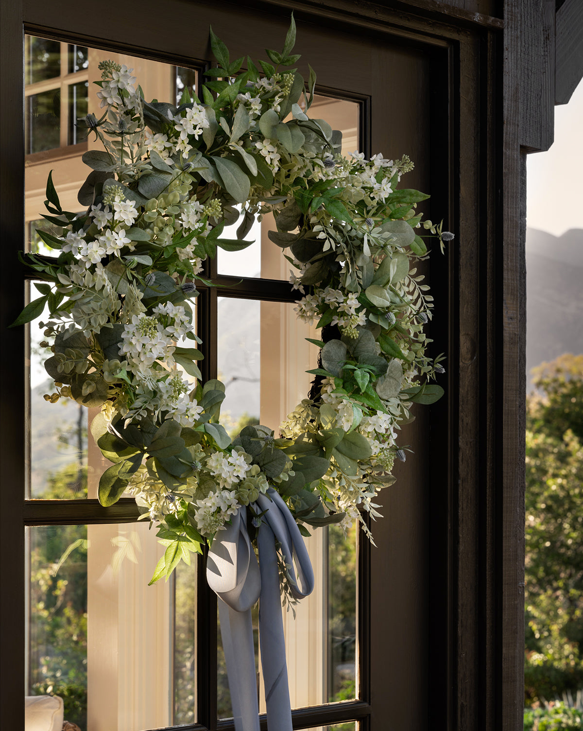 An Orange Jasmine & Eucalyptus Wreath with green and white foliage and a blue ribbon hangs on a glass door, reflecting trees and mountains outside.