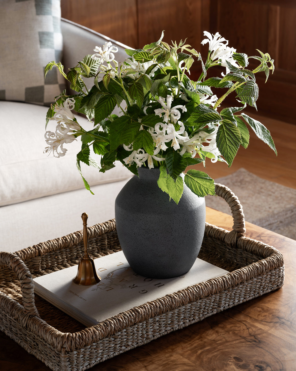 A woven tray on a wooden table holds the Delgado Vase with white flowers and green leaves, a small gold bell, and a closed book. In the background, there’s a beige sofa with a patterned pillow, by McGee & Co.