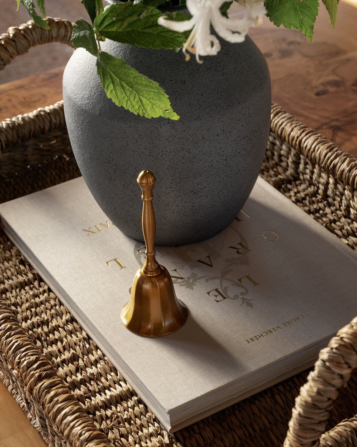 The Elsher Bell, crafted in golden antique brass, sits atop a large gray book with gold lettering beside a gray vase of white flowers and green leaves, all arranged in a woven tray for classic vintage decor, by McGee & Co.