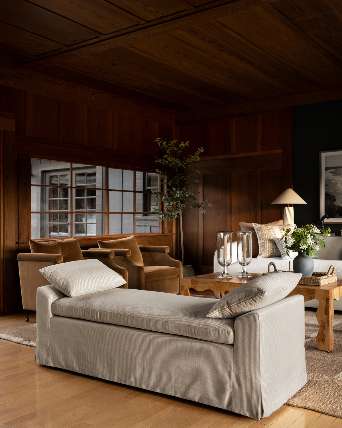 A cozy living room with wooden walls and ceiling features a beige daybed, two brown velvet armchairs, the Scaletta Bench (Ready to Ship) with linen ticking stripe upholstery, a wooden coffee table with candles and flowers, and large windows by McGee & Co.