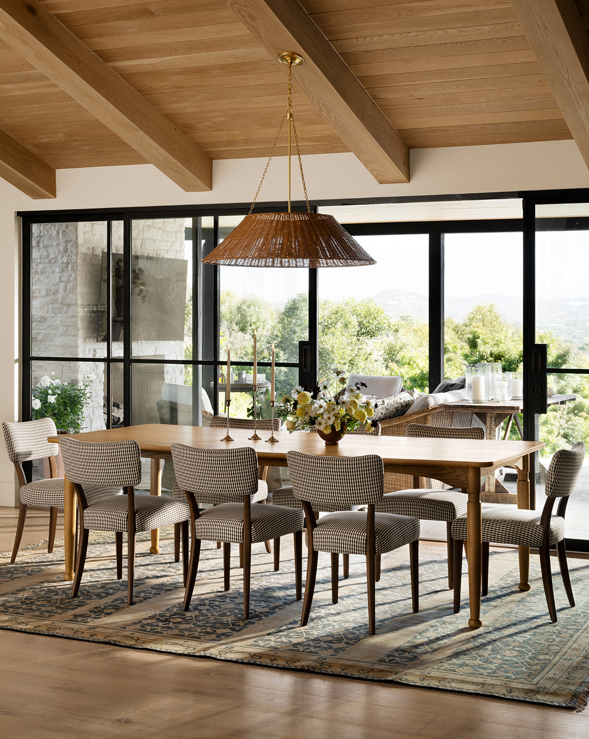 A dining room featuring the handcrafted Phipps Dining Table, eight upholstered chairs, a woven pendant light, wood-beam ceilings, a patterned rug, and large glass doors opening to a patio with greenery by McGee & Co.