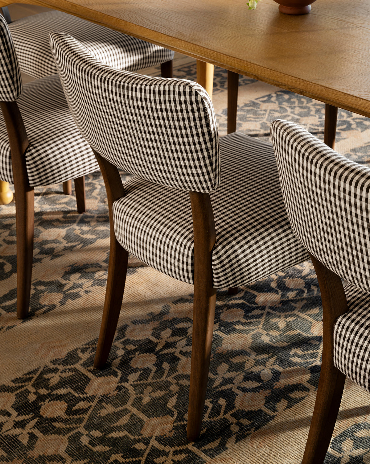 A close-up of Makerspalm Etienne Dining Chairs, featuring wooden legs and black-and-white checkered upholstery, arranged by a wooden dining table on a patterned carpet. McGee & Co.