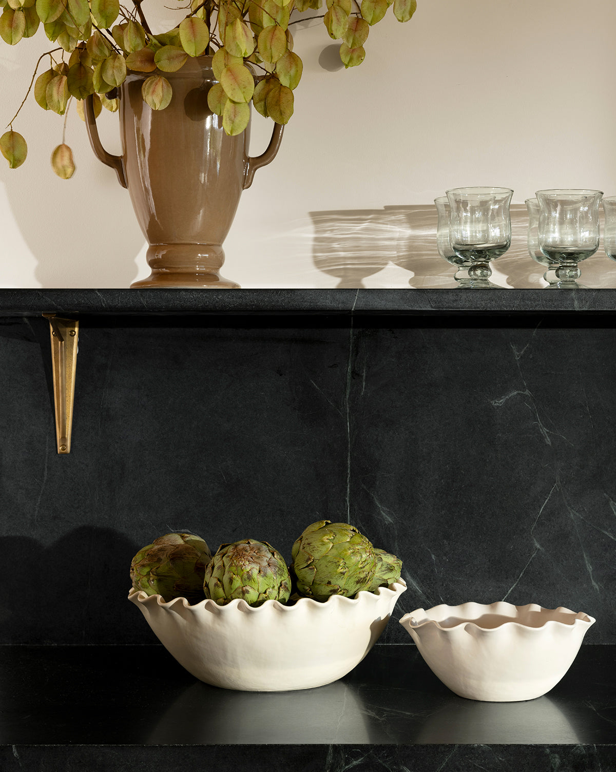 A dark marble shelf displays the Olindo Serving Bowls (Set of 2), one filled with artichokes. Above, a beige vase with dried leaves and two clear glasses are arranged against a pale wall, by McGee & Co.