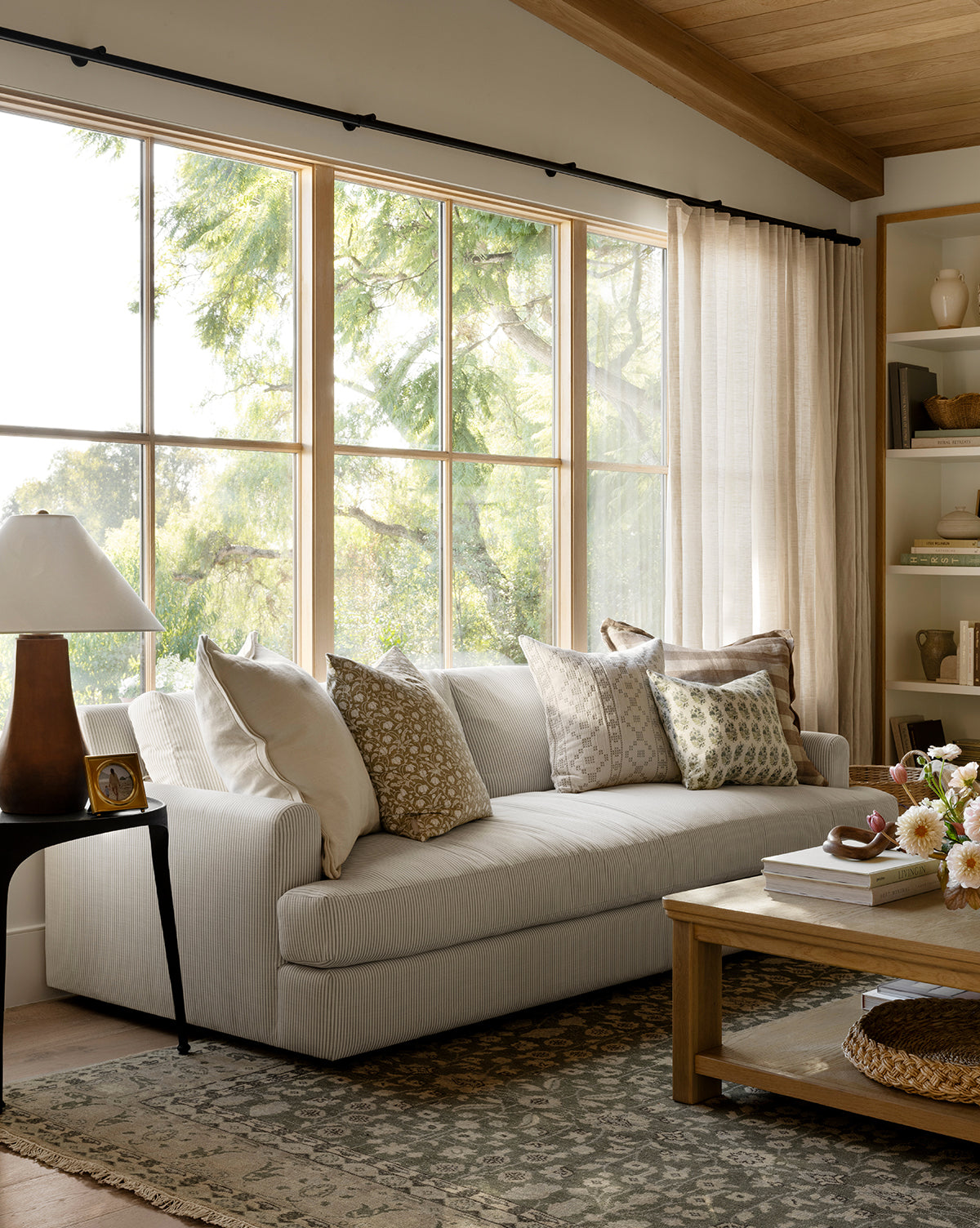 A bright living room features the Makerspalm Winthrop Sofa 90 with patterned pillows, a wooden coffee table with flowers, a side table and lamp, built-in decor shelves, light curtains, and a patterned rug for a cozy finish from McGee & Co.