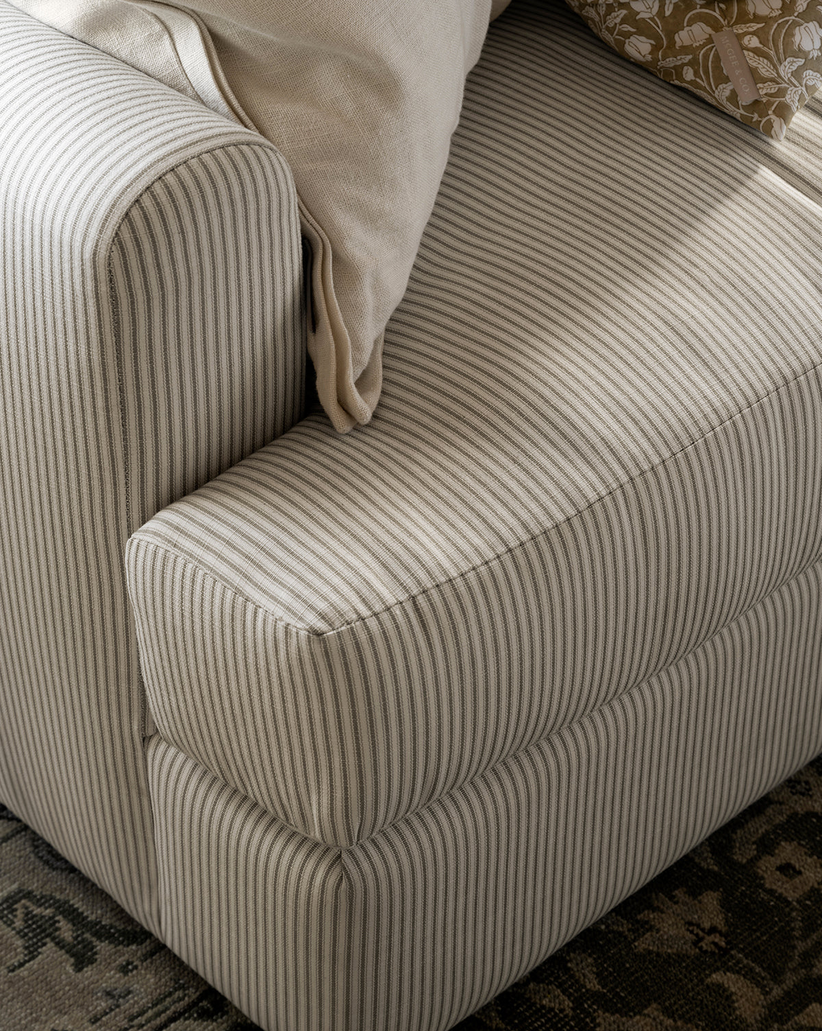 Close-up of the Winthrop Sofa 90 in beige and gray stripes, featuring a light cream pillow and a floral-patterned pillow on the armrest, set beside a dark patterned rug, by McGee & Co.