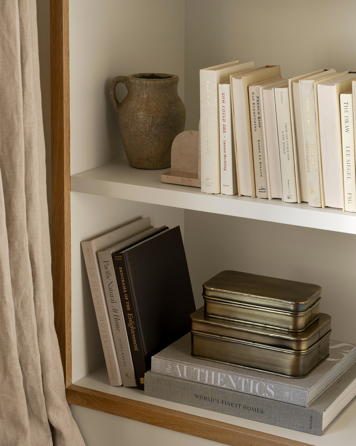 A bookshelf holds neutral-toned books, two stacked metal tins, a small arch decor piece, large hardcover architecture books, and features the Sabine Pitcher Vase. Beige curtains hang to the side, by McGee & Co.