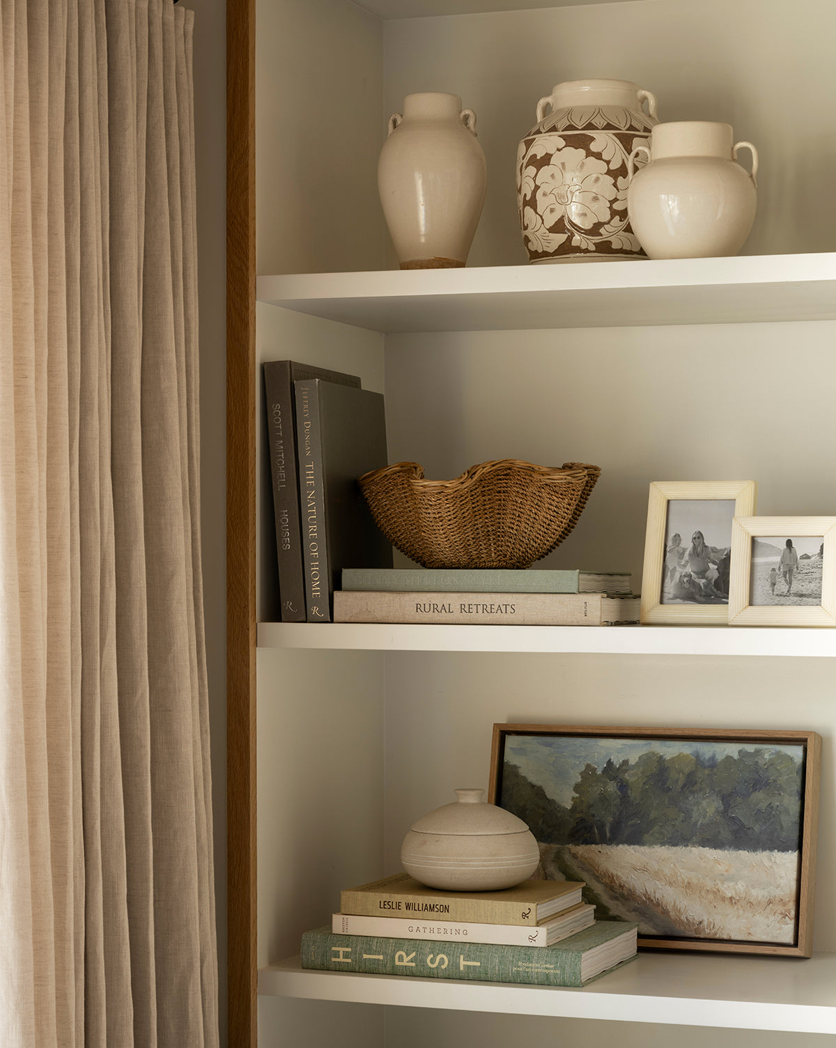 A styled bookshelf with the Blaise Vase, cream vases, a woven basket, stacked books, framed black-and-white photos, and a landscape painting sits next to beige curtains. The home decor features neutral and natural tones by McGee & Co.