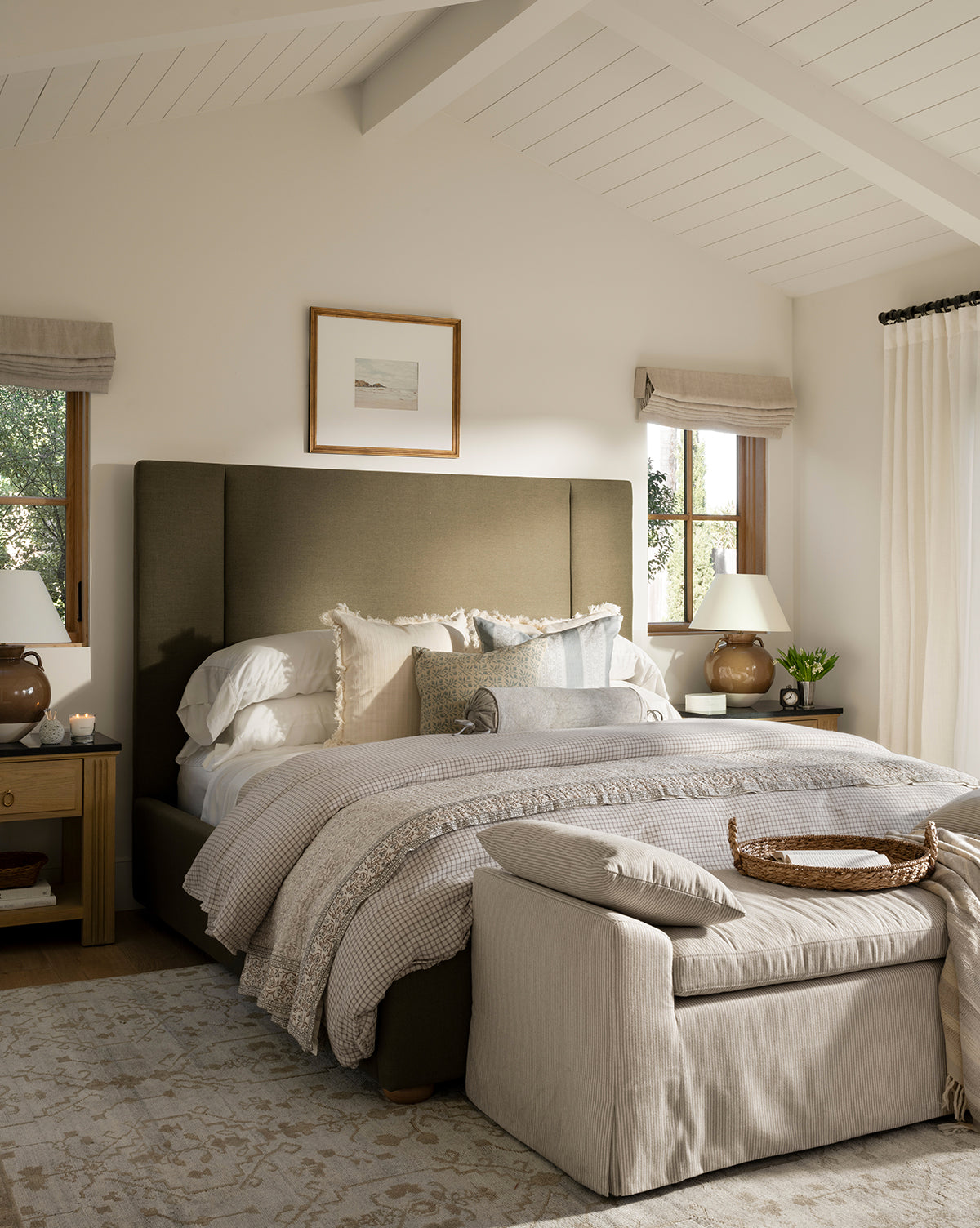 A cozy bedroom features a plush bed with neutral bedding, pillows, and a soft throw. The Lexington Hand-Knotted Wool Rug anchors the space, paired with matching nightstands, lamps, art above the bed, and plenty of natural light, by McGee & Co.