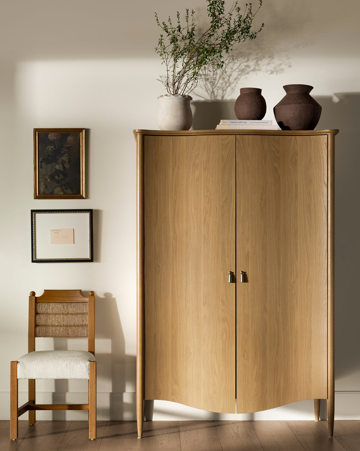 A curved wooden cabinet stands against a light wall. On top sit two brown vases, a book, and the Frederick Paper Mache Vase with green branches. A wooden chair is beside it and three framed pictures hang on the wall, by McGee & Co.
