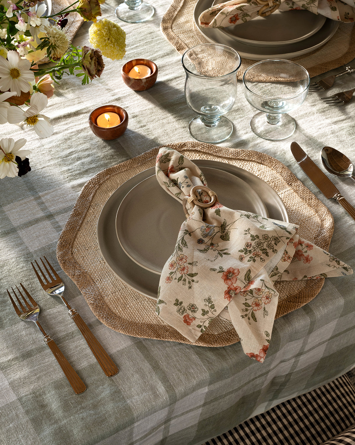 A beautifully set table with woven placemats, Aiden Soft Gray Side Plates, floral napkins with rings, glassware, wooden-handled cutlery, tealight candles, and a vase of flowers on a plaid tablecloth, by McGee & Co.