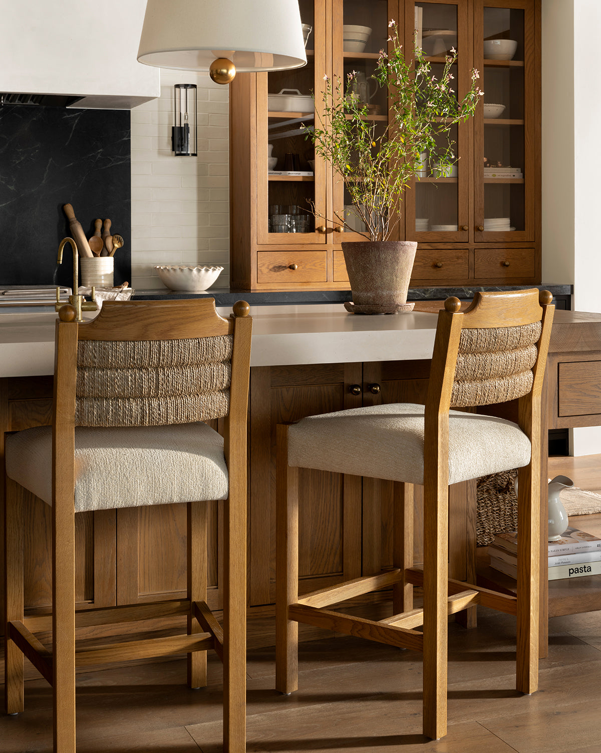 A cozy kitchen features honey oak cabinets, a white countertop, two Makerspalm Alberta Counter Stools with woven seagrass backs, a potted plant on the island, and shelves stocked with dishes and books in the background—McGee & Co.