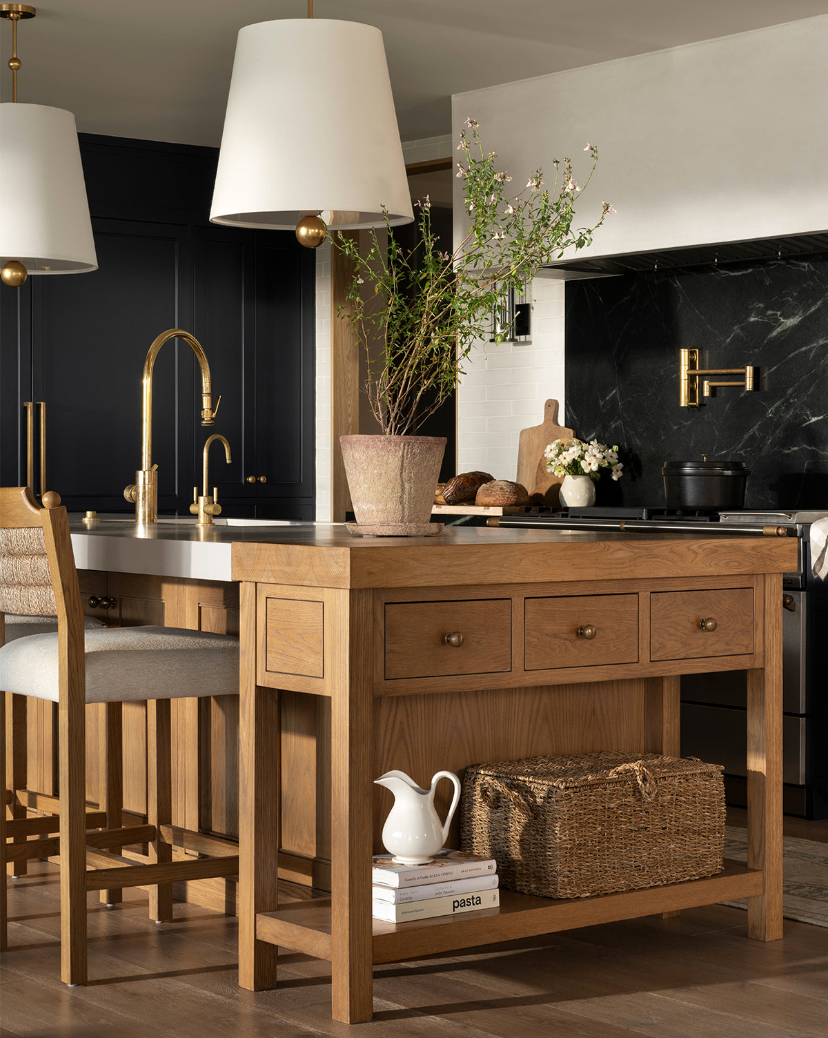 A modern kitchen features a wooden island, gold fixtures, a potted plant, the Kya Seagrass Box, a white pitcher and pasta jar. Large pendant lights hang above dark cabinetry and a marble backsplash in the background, by McGee & Co.