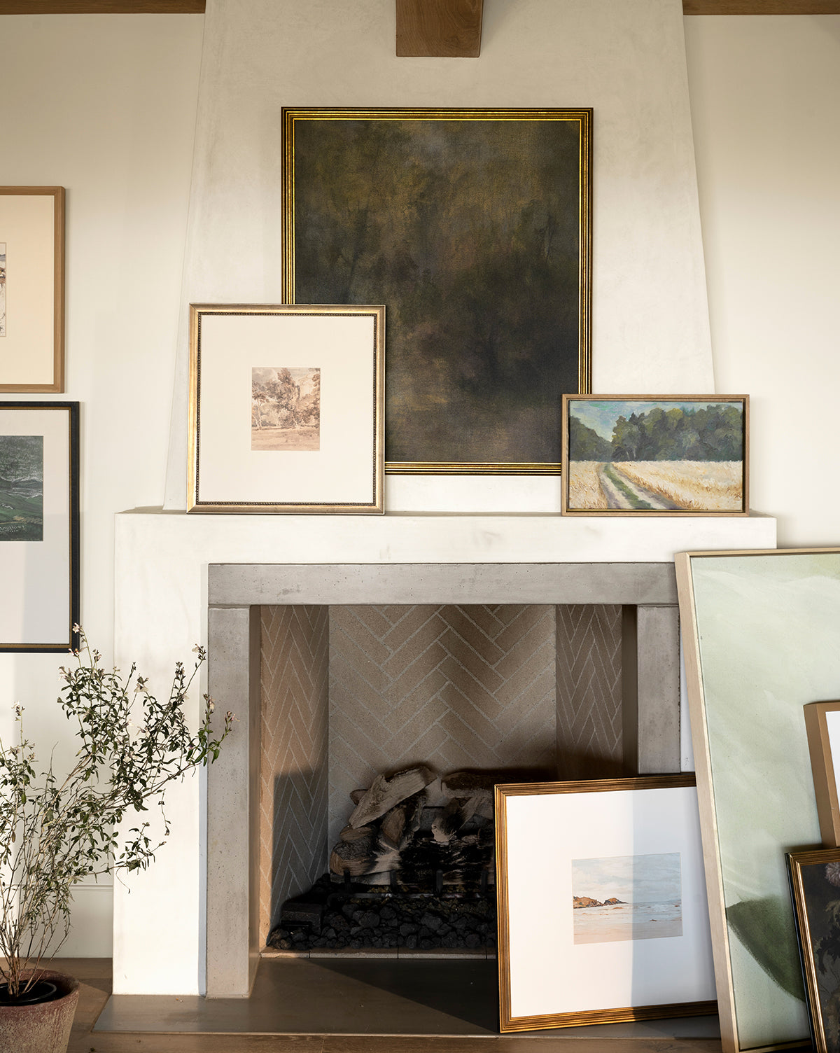 A modern white herringbone fireplace showcases Dream State, a museum-quality canvas landscape painting, framed and displayed on the mantel and floor. Expressive artwork complements the scene, with a potted plant to the left, by McGee & Co.